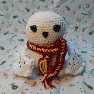 Peut inclure: Peluche hibou en crochet blanc portant une écharpe rouge et jaune. Le hibou a des yeux noirs et un bec noir.