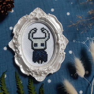 Peut inclure: Un cadre ovale blanc avec une finition argentée, contenant une broderie au point de croix d'un chevalier bleu et noir. Le chevalier a un visage blanc et de grandes oreilles pointues. Le fond de la broderie au point de croix est blanc.