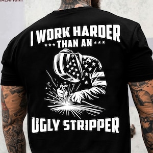 Op de afbeelding: Zwart T-shirt met een witte afbeelding van een lasser en de tekst "I WORK HARDER THAN AN UGLY STRIPPER". De helm van de lasser heeft een sterrenpatroon. Het shirt heeft een ronde hals.