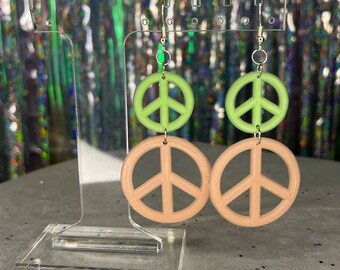 May include: A pair of dangle earrings with a peach and green peace sign design. The earrings are on a clear acrylic stand.