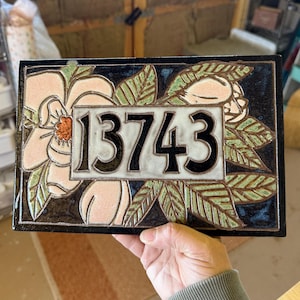 May include: A rectangular ceramic house number plaque featuring the digits 13743 in black. The design includes a cream-colored central panel surrounded by a dark blue border, decorated with light pink flowers and green leaves with brown outlines.