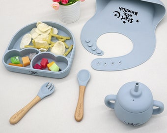 May include: A blue silicone baby bib with the text "Start with Bismillah" and a crescent moon. A blue silicone divided plate with a spoon and fork. A blue silicone sippy cup with a straw.