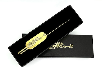 May include: A gold bookmark with Arabic calligraphy in black. The bookmark is in a black box with the same calligraphy on the box.