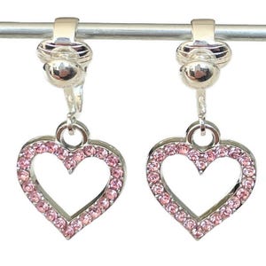 Puede incluir: Un par de pendientes de clip plateados con dijes de corazón de cristal rosa. Los corazones tienen un centro hueco y están rodeados de pequeños cristales rosas.