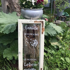 May include: A white window frame with a glass pane featuring a dandelion design and the text "Genie&szlig;e die kleinen Dinge Sie machen das Leben gro&szlig;artig". The window frame is mounted on a wooden post and a metal planter with white and purple flowers sits on top of the post.