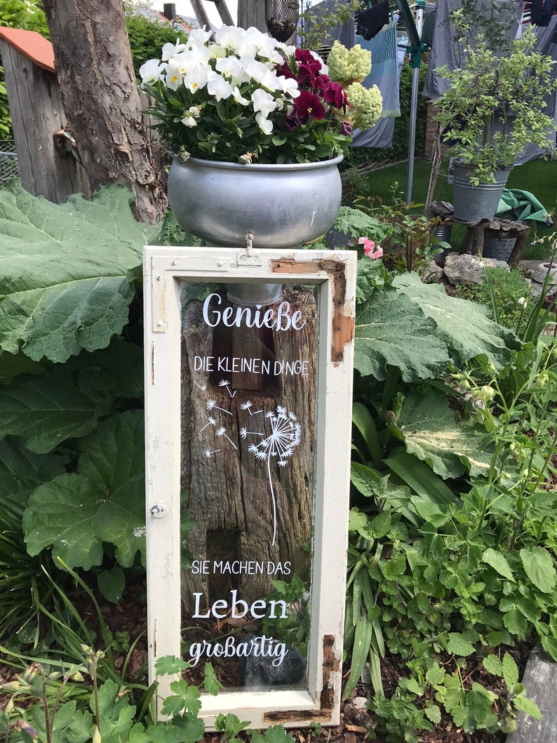 May include: A white window frame with a glass pane featuring a dandelion design and the text "Genie&szlig;e die kleinen Dinge Sie machen das Leben gro&szlig;artig". The window frame is mounted on a wooden post and a metal planter with white and purple flowers sits on top of the post.