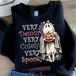 May include: Black sweatshirt with a white ghost holding a jack-o'-lantern. The text on the shirt reads "Very Demure Very Cutesy Very Spooky".