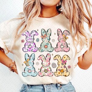 May include: A white t-shirt with six floral bunny rabbits printed on the front. The bunnies are in different colors and have a pastel color scheme. Each bunny has a bow and is surrounded by small flowers.