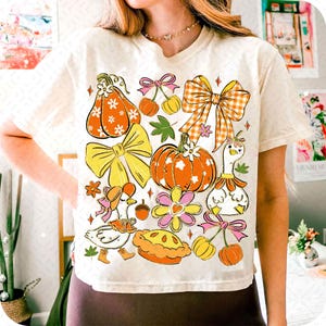 May include: Cream-colored t-shirt featuring a colorful fall-themed graphic. The design includes pumpkins, bows, flowers, cherries, and a goose wearing a hat. The graphic is in shades of orange, yellow, pink, and green.