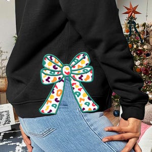 Puede incluir: Sudadera negra con un gran lazo colorido aplicado. El lazo presenta un fondo blanco con formas naranjas, amarillas, verdes y moradas, delineadas en verde. La persona lleva vaqueros azules.