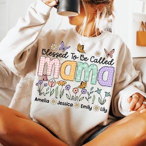 Puede incluir: Una sudadera blanca con un dise&ntilde;o floral y el texto "Blessed To Be Called Mama" con los nombres Amelia, Jessica, Emily y Lily debajo.