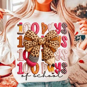 Pode incluir: Camisola branca com o texto "100 Days of School". O design inclui um la&ccedil;o com estampa de leopardo, l&aacute;pis e n&uacute;meros em v&aacute;rias cores. O texto est&aacute; em um estilo l&uacute;dico e manuscrito, perfeito para celebrar um marco escolar.