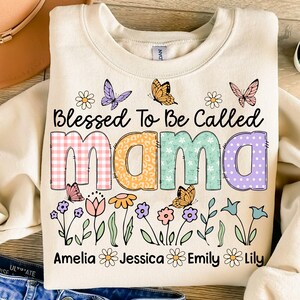 Puede incluir: Una sudadera beige con el texto "Blessed To Be Called Mama" en letras de colores con patrones. Las letras est&aacute;n decoradas con cuadros, estampado de leopardo, lunares y motivos florales. Debajo del texto hay una fila de flores y mariposas. Los nombres Amelia, Jessica, Emily y Lily est&aacute;n escritos debajo de las flores.