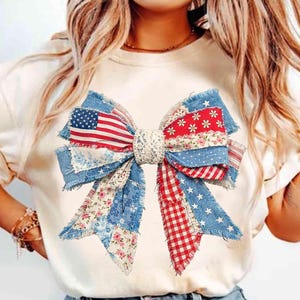 May include: A cream-colored t-shirt featuring a large, patriotic bow design. The bow is crafted from various fabrics, including denim, floral prints, and a section resembling the American flag. The bow has a lace center and frayed edges.