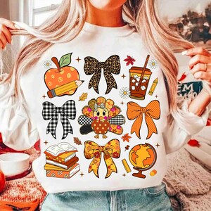 May include: White long-sleeved sweatshirt featuring a collection of fall-themed graphics. Includes an apple with a pencil, bows, a turkey, books, a globe, and a coffee cup. The graphics are in various colors and patterns.