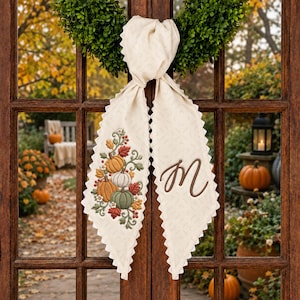 May include: A cream-colored fabric door hanger featuring a scalloped edge and a tied top. The left tail displays an embroidered design of pumpkins, leaves, and berries. The right tail features a large, brown, cursive letter M monogram.