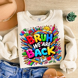 Puede incluir: Una sudadera beige con un estampado gr&aacute;fico colorido que dice "Bruh We Are Back" rodeado de &uacute;tiles escolares como l&aacute;pices, crayones y un arco&iacute;ris.