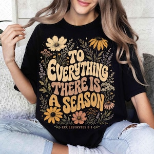 Puede incluir: Una camiseta negra de manga corta con un estampado gráfico que incluye el texto "TO EVERYTHING THERE IS A SEASON" y "ECCLESIASTES 3:1" en una fuente ondulada, rodeado de ilustraciones de varias flores silvestres en tonos amarillo, naranja y marrón.