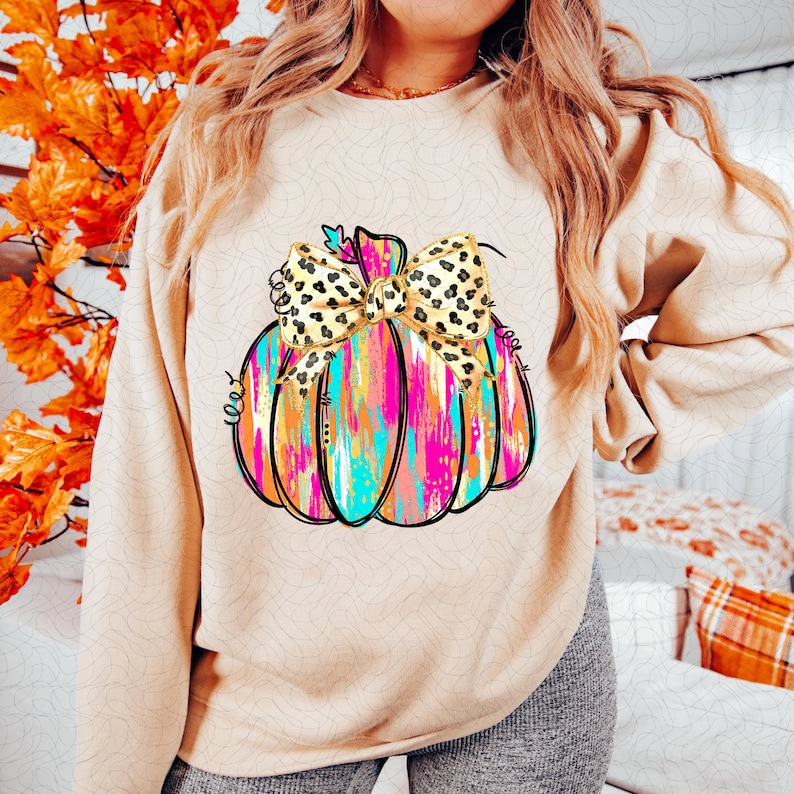 May include: A beige sweatshirt with a colorful abstract pumpkin design. The pumpkin has a large yellow and black leopard print bow on top.