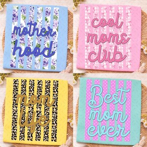 May include: Four square decorative canvases with text. The top left canvas has the words "mother hood" in blue over a floral and striped background. The top right canvas has the words "cool moms club" in pink. The bottom left canvas has the words "mom life" in gold. The bottom right canvas has the words "Best mom ever" in blue.