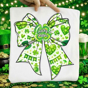 May include: White t-shirt featuring a large green bow design with shamrocks, hearts, and banners. The bow has a black outline and the words "Lucky charm" in the center. The design is perfect for St. Patrick's Day.