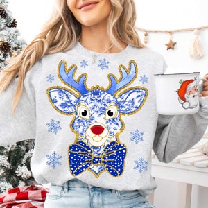 Op de afbeelding: Een grijs sweatshirt met een blauwe en gouden rendier dat een blauwe en witte strik draagt. Het rendier heeft een rode neus en is omringd door sneeuwvlokken.