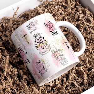 May include: White ceramic mug with a pink and gold design. The mug features inspirational quotes and bible verses, including "Be Still and Know that I am God", "God is good all the time", "Heal me O Lord", and "Pray Big Win Small".