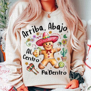 以下が含まれることがあります： ベージュのトレーナーに、黒い文字で「Arriba. Abajo. Al Centro Pa'Dentro」と書かれています。ソンブレロをかぶったジンジャーマンがオレンジジュースの入ったグラスを持っています。マラカスやその他の華やかな装飾があります。