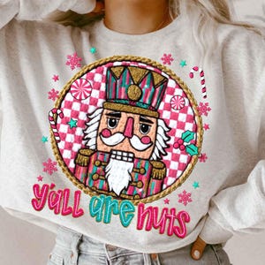 Könnte beinhalten: Ein hellgraues Sweatshirt mit einem farbenfrohen Grafikdesign eines Nussknackers und dem Text "Y'all are nuts." Das Design umfasst rosa, türkis, goldene und weiße Elemente, mit Zuckerstangen und Schneeflocken. Ein festlicher Artikel.
