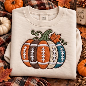 May include: A cream-colored crewneck sweatshirt featuring an embroidered graphic of a pumpkin composed of five vertical segments, each resembling a football with white laces. The design includes a green stem and an orange leaf on top.