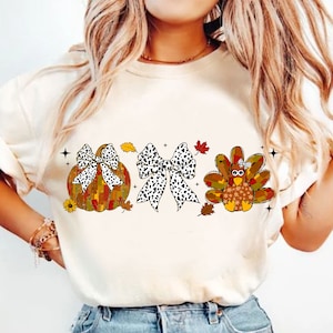 May include: Cream-colored t-shirt featuring a fall-themed graphic. The design includes a pumpkin, a bow, and a turkey, all adorned with a black and white spotted pattern and autumn leaves. The shirt is a casual wear item.