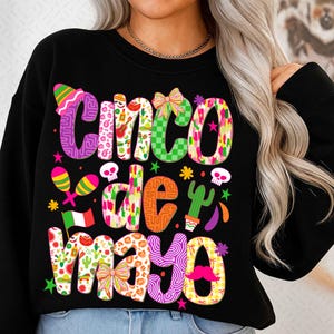 Può includere: Felpa nera con un colorato design "Cinco de Mayo". Il testo è multicolore e fantasia, con elementi come un sombrero, maracas e un cactus. Il design è festoso e celebra la festa messicana.