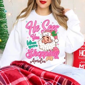 May include: White Christmas sweatshirt with a pink graphic of Santa Claus and the text "He Sees You When You're Shopping Amelia".