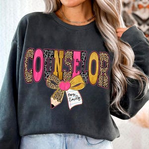 May include: Dark grey sweatshirt featuring the word "COUNSELOR" in pink, gold, and leopard print. A pencil, apple, and bow graphic is below the word. The name "Mrs. Emily" is written on a small notepad.