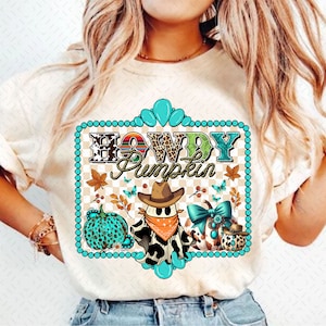 Op de afbeelding: Een wit T-shirt met een turquoise frame en de tekst "Howdy Pumpkin" in kleurrijke letters. Binnen het frame is een geest die een cowboyhoed en bandana draagt, en twee pompoenen met turquoise en bruine accenten.