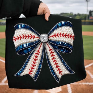 May include: A black decorative bow with a baseball theme. The bow is embellished with white pearls, red stitching, and blue gems. A large, square, clear gem is in the center of the bow. The background is a baseball field.
