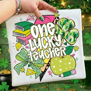May include: White graphic tee with the text "ONE LUCKY TEACHER" in a playful font. The design includes a green apple, shamrocks, books, and a bow. The tee is held up by a hand, showcasing the design's vibrant colors and St. Patrick's Day theme.