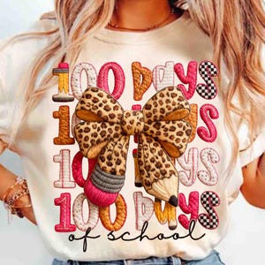 Pode incluir: Camiseta creme com um gr&aacute;fico colorido celebrando 100 dias de escola. O design apresenta o texto "100 dias de escola" com um la&ccedil;o com estampa de leopardo e detalhes de l&aacute;pis. Os n&uacute;meros e letras s&atilde;o de v&aacute;rias cores.