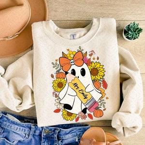 May include: A cream colored sweatshirt with a graphic of a ghost holding a pencil. The ghost is wearing a bow and surrounded by fall leaves and sunflowers. The text "Mrs. Emily" is written on the pencil.
