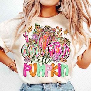 Puede incluir: Camiseta color crema con un gráfico colorido de tres calabazas con detalles florales y de hojas. Las calabazas están decoradas con patrones rosas, naranjas y turquesas. El texto "hello PUMPKIN" está debajo.