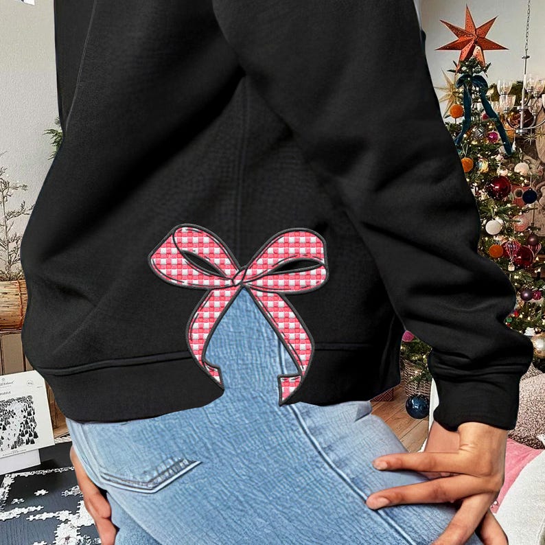 Puede incluir: Sudadera negra con un lazo de cuadros rojos y blancos en la espalda. La sudadera se lleva sobre unos vaqueros azules.