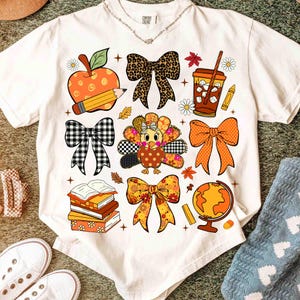 May include: White t-shirt featuring colorful fall-themed graphics. The design includes an apple with a pencil, bows, a turkey, books, a globe, and a coffee cup. The shirt has a crew neck and short sleeves.