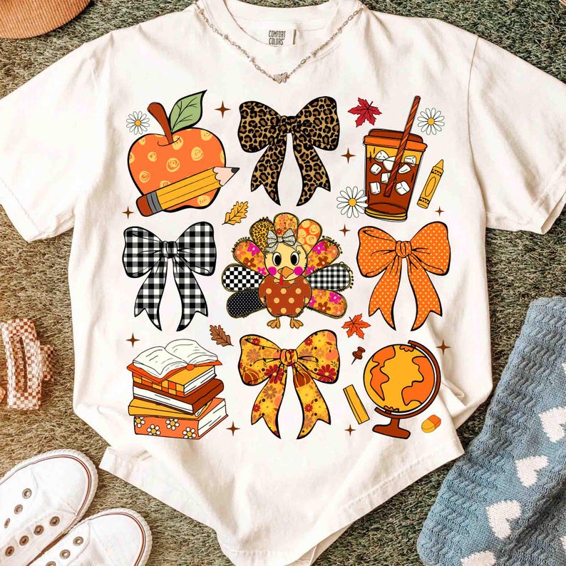 May include: White t-shirt featuring colorful fall-themed graphics. The design includes an apple with a pencil, bows, a turkey, books, a globe, and a coffee cup. The shirt has a crew neck and short sleeves.