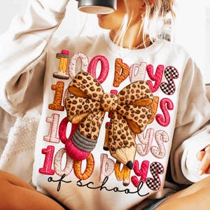 Pode incluir: Camisola branca com um design bordado colorido. O design apresenta o texto "100 Days of School" com um grande la&ccedil;o com estampa de leopardo e detalhes de l&aacute;pis. Os n&uacute;meros e as letras s&atilde;o de v&aacute;rias cores.