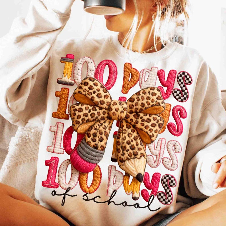 Pode incluir: Camisola branca com um design bordado colorido. O design apresenta o texto "100 Days of School" com um grande la&ccedil;o com estampa de leopardo e detalhes de l&aacute;pis. Os n&uacute;meros e as letras s&atilde;o de v&aacute;rias cores.