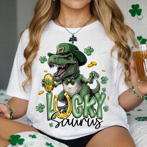 May include: A white t-shirt with a green dinosaur wearing a green hat and holding a gold coin. The dinosaur is surrounded by shamrocks and the text "Lucky Saurus".