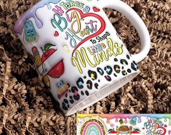Puede incluir: Taza de café de cerámica blanca con un diseño colorido que presenta el texto "Se necesita un gran corazón para moldear las mentes pequeñas". La taza tiene un patrón de estampado de leopardo blanco y negro alrededor de la parte inferior. La taza está sentada en una trituradora de papel marrón.