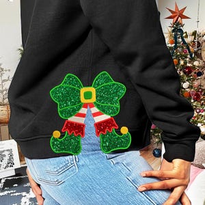 Puede incluir: Sudadera negra con un lazo de zapato de elfo de purpurina verde en la espalda. La sudadera la lleva puesta una persona con vaqueros azules.