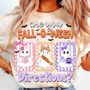 May include: Beige t-shirt with the text "Are you Fall-O-Ween Directions?" and three ghost illustrations. Each ghost has a different accessory: a book, a pencil, and a jack-o'-lantern. The design includes bats and decorative frames.