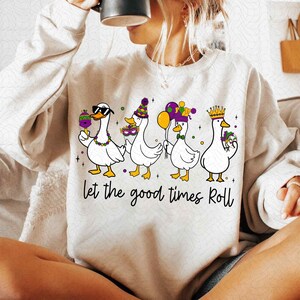 May include: Cream-colored sweatshirt featuring a graphic of four white ducks dressed for a celebration. Each duck is wearing festive accessories like sunglasses, hats, and crowns, holding items like a lollipop and balloons. The phrase "Let the good times Roll" is written below.